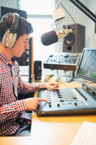 Radio host wearing headphones operating sound mixer on table in studio